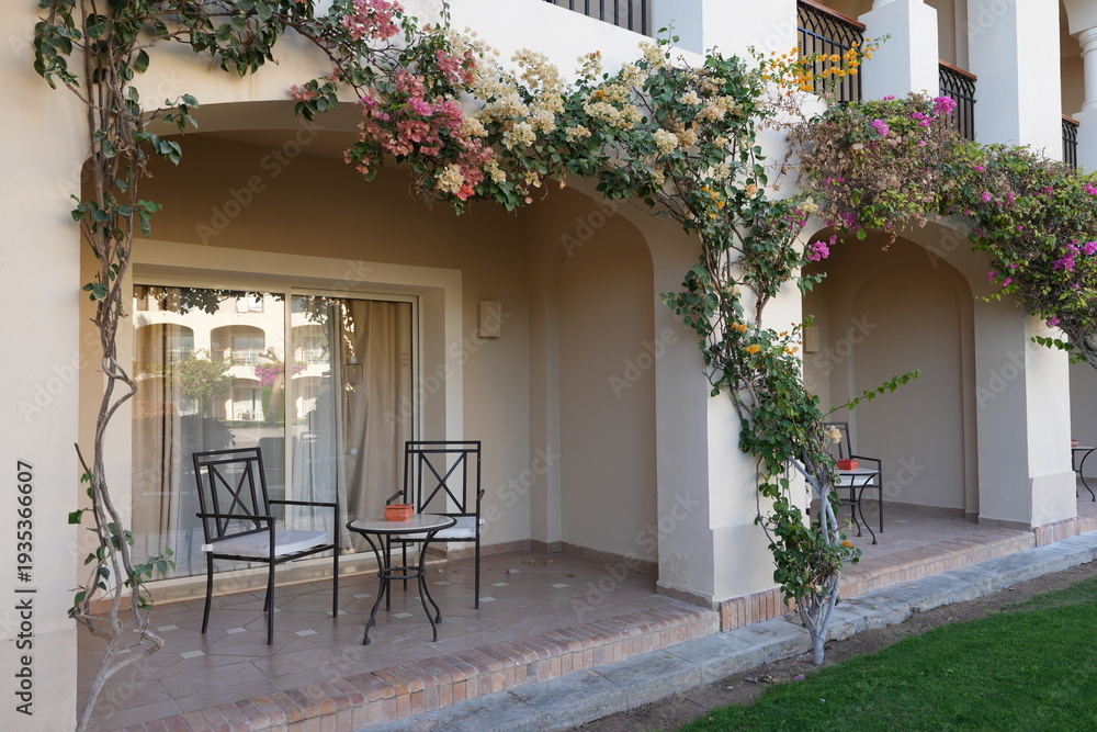 Obraz premium Outdoor patio area with two black metal chairs and a small table adorned with flowers and greenery, featuring a glass door leading to an interior space