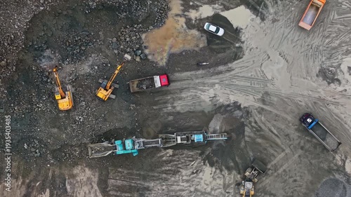 Workers operate machines and vehicles at a granite quarry site. Heavy equipment digs and transports stones. Dust and rocky ground cover the area.