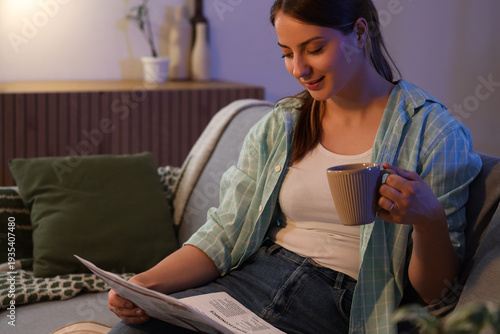 Young woman with cup of tea reading newspaper on sofa at home in evening