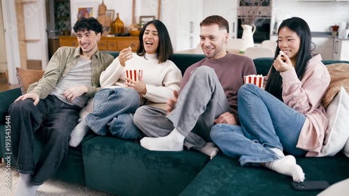 Cheerful group of young friends laughing and eating popcorn while watching a movie on the sofa in the living room. Cozy home entertainment