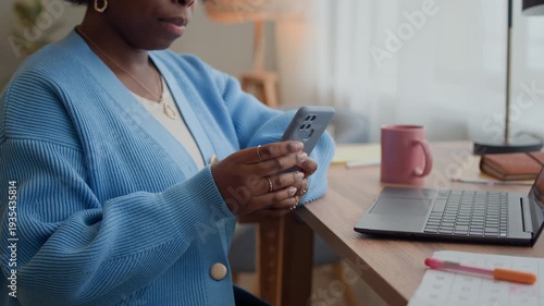 Cropped shot of young Black woman highlighting dates in calendar while checking smartphone, sitting at home table with laptop