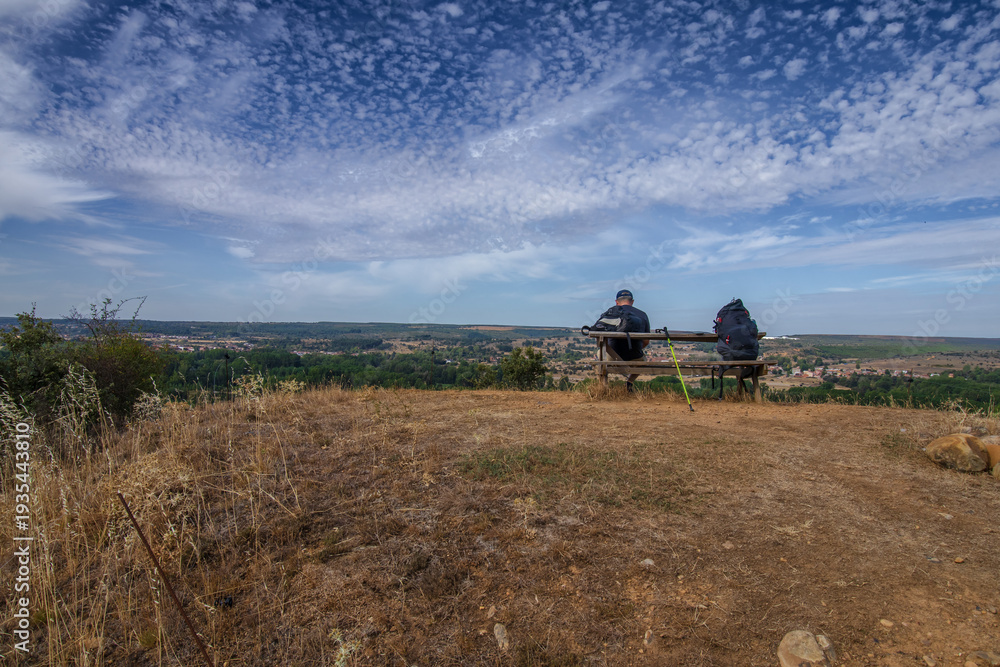 Fototapeta premium Un caminante descansa en un banco de madera junto a su mochila, contemplando un vasto paisaje rural bajo un cielo de nubes dispersas, capturando la esencia del Camino de Santiago