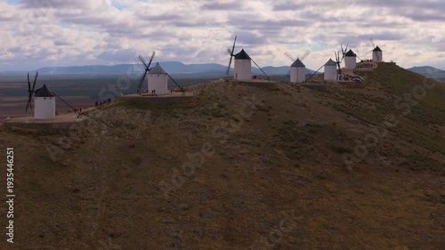 Aerial view of a line of iconic white windmills atop a hill with people walking around, creating a picturesque scene, Consuegra, Castile-La Mancha, Spain.
