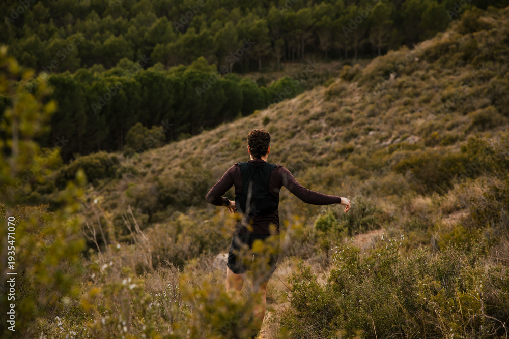 Obraz premium Rear view of a trail runner moving along a rugged mountain trail surrounded by wild nature and forested hills. Outdoor adventure sport and endurance lifestyle concept.