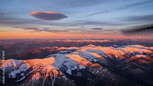 Snow-covered mountain range at sunset, aerial view with orange sky and lenticular cloud
