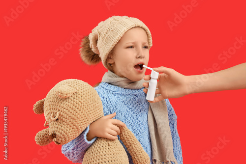 Sick little girl with toy bear and woman spraying her sore throat on red back...