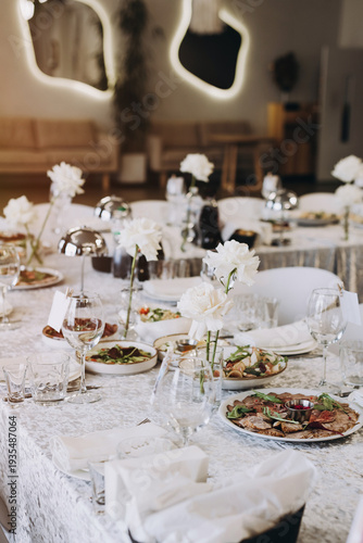 Elegant banquet table setting with fresh white roses, cordless silver lamps, and gourmet appetizers in a modern high-end restaurant interior