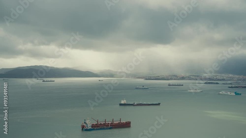 Multiple cargo ships and ferries anchored in the Bay of Gibraltar as dramatic storm clouds roll over the coastline.
