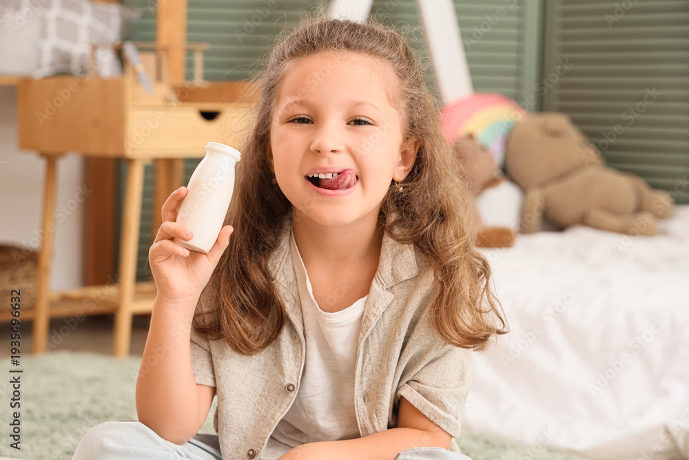 Fototapeta premium Cute little girl with bottle of yogurt showing tongue in room
