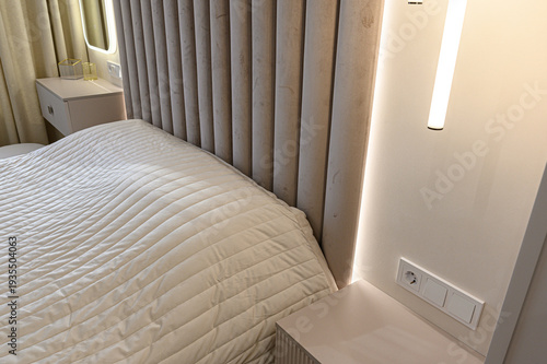 A modern bedroom with a quilted beige bed, plush ribbed headboard, sleek white nightstand, built-in LED lighting, and minimalist decor