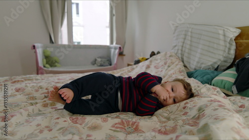 Baby lying on bed stretching arms with drowsy expression, relaxed and sleepy, gentle childhood moment in cozy family bedroom with soft bedding and natural light