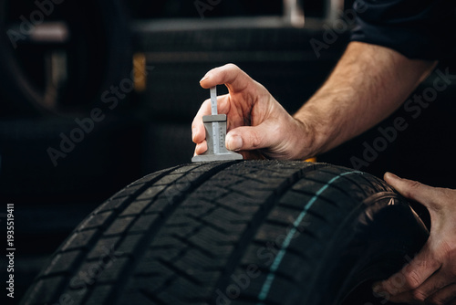 Hand of a technician measuring tire tread depth with a gauge on a black tire in a workshop surrounded by additional tires in the background