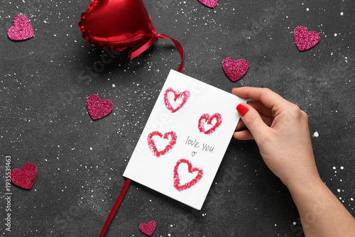 Female hand with red manicure, festive postcard and decor on black grunge background. Valentine's Day celebration