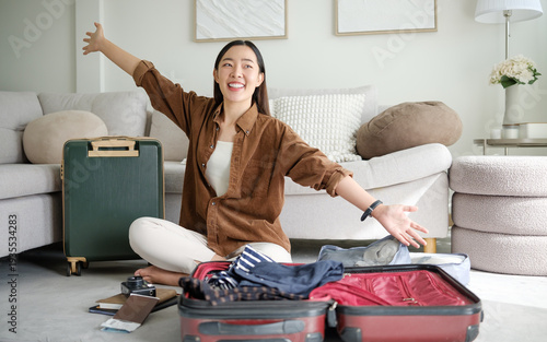 Excited young woman packing suitcase at home with arms wide open. Travel preparation and vacation planning concept.