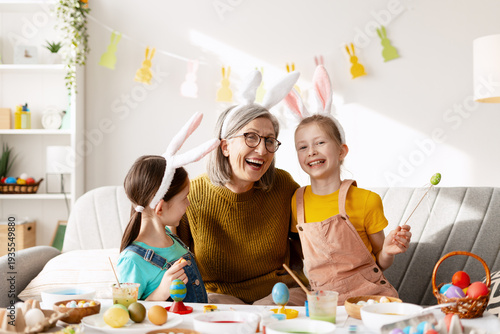 Grandparent and two granddaughters smiling, painting Easter eggs in a decorated home