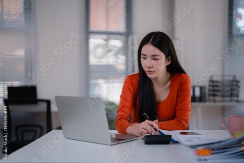 Young Asian woman concentrating on crunching numbers and checking financial documents while calculating expenses on her laptop in a modern office setup