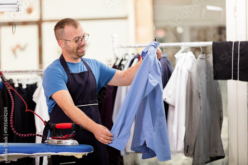 Male laundry worker checking cleaned garments on a rack in a professional dry cleaning store.