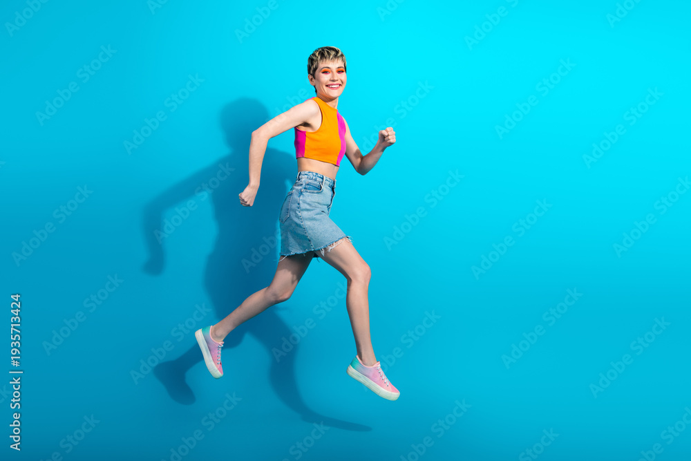 Obraz premium Young woman jumps in colorful top and denim skirt against blue background for a vibrant fashion lifestyle stock photo
