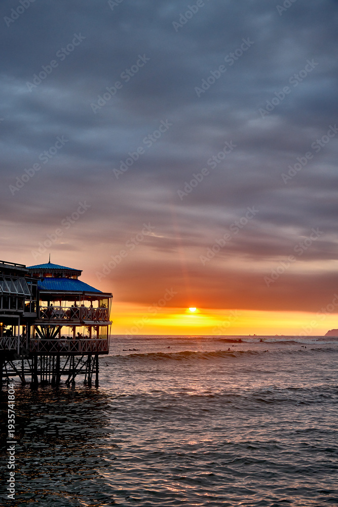 Fototapeta premium La Rosa Náutica glows above the Pacific at sunset in Miraflores, Lima, as surfers and evening visitors embrace an active coastal lifestyle, reflecting safe travel experiences and the growing appeal of