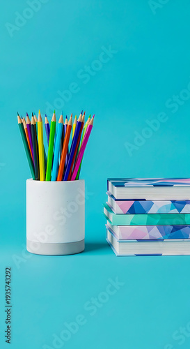 Colorful pencils in a white cup next to stacked notebooks on blue background