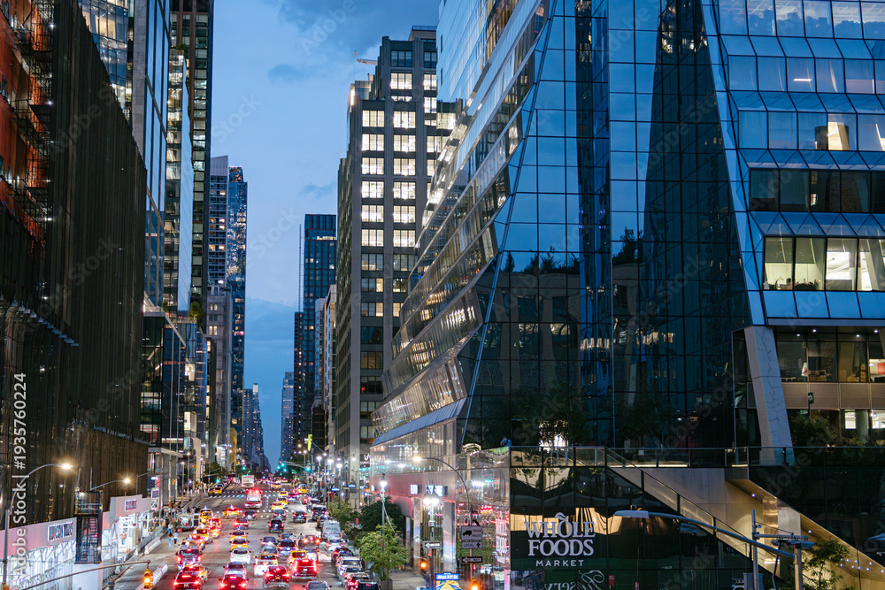 Fototapeta premium New York City, New York, USA - 08.02.2024: Evening traffic moves through Hudson Yards in Manhattan with illuminated glass skyscrapers and Whole Foods Market visible. The night city scene captures