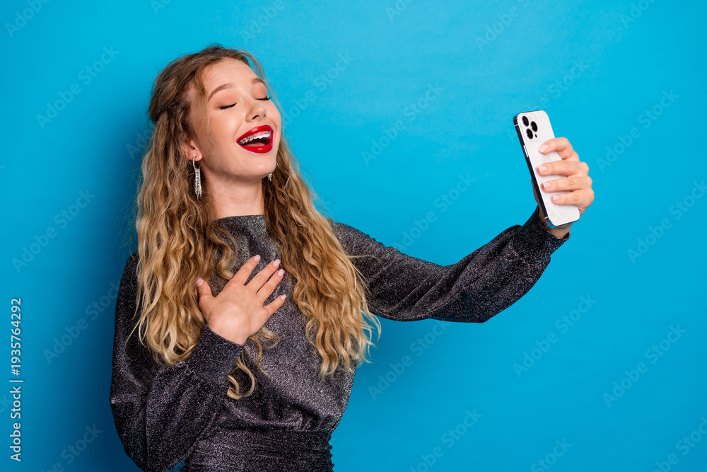 Obraz premium Joyful girl in a shiny dress takes a selfie against blue background showcasing glamour confidence fashion lifestyle social media stock