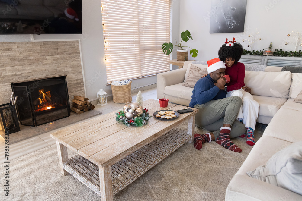 Naklejka premium African American couple lounging in living room on sofa by fireplace wearing Santa hats, copy space