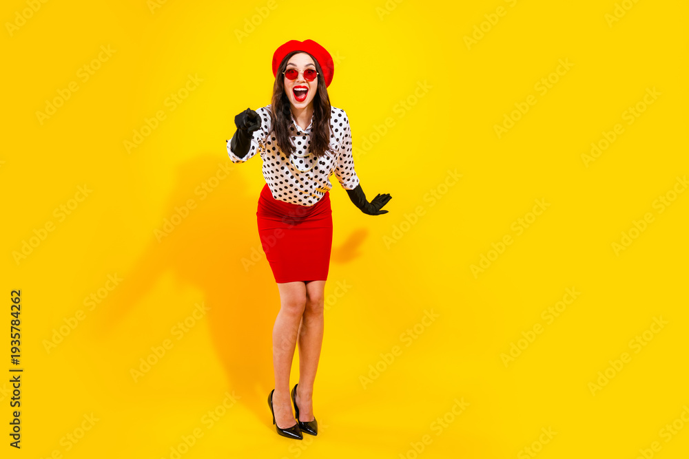 Obraz premium Happy young woman in red beret and pencil skirt wearing polka dot blouse, black gloves and red sunglasses posing and pointing on a bright yellow background in retro fashion style