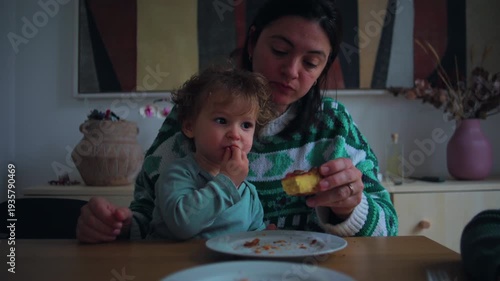 Mother eating piece of cake while toddler sits beside her at table, candid family snack time moment expressing warmth, appetite and everyday motherhood