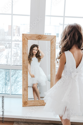 Fotografie A little girl in a white dress looks at her reflection in the mirror, childish dreaminess and self-perception