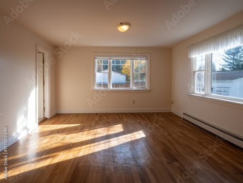 Sunlight streams through windows onto hardwood floors. Room features clean walls and a central ceiling light. Natural light illuminates empty space with warm tones. Windows offer views of trees