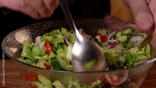 The cook stirs the salad with tomatoes, olives and green lettuce leaves in a bowl with a spoon.