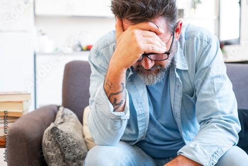 Картината върху платно Sad and frustrated 50s man suffering from stress, depression at home, sitting on sofa, touching head, feeling headache, migraine, dealing with loss, grief, trauma