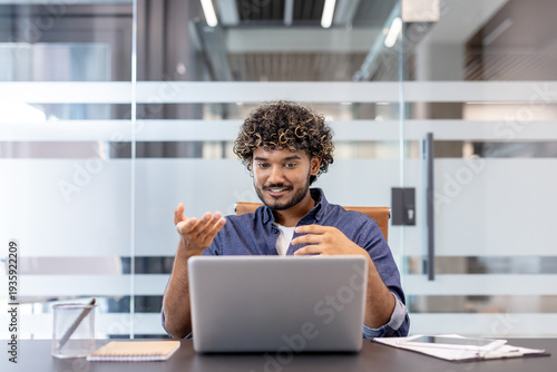 Young Indian man sitting at a desk in an office with a laptop and talking on a video call with stiff hands