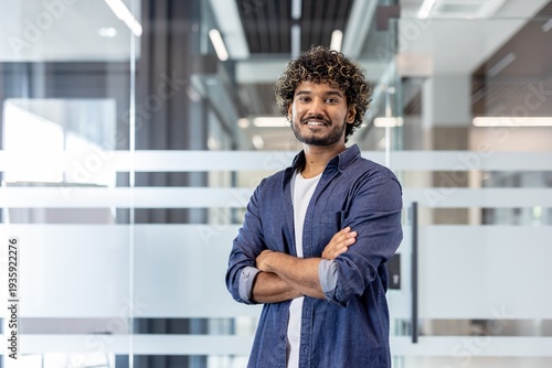 Portrait of a young smiling Indian man in a shirt standing in the office, arms crossed on his chest and looking confidently at the camera