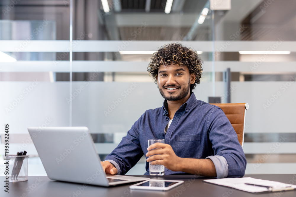 Fototapeta premium Portrait of a young Indian man sitting at a desk with a laptop, holding a glass of water in his hand and looking at the camera with a smile