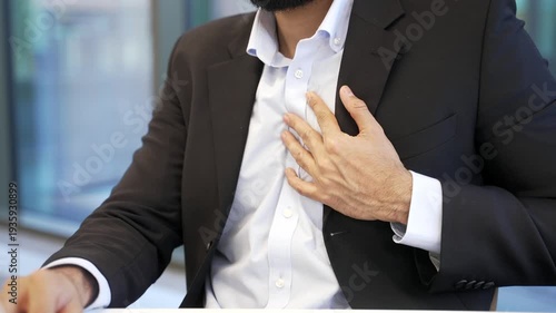 Close up of man's hands holding massaging chest. Businessman in formal suit has heart attack while sitting at a workplace in business office. Unwell man worker is breathing deeply needs medical help. 