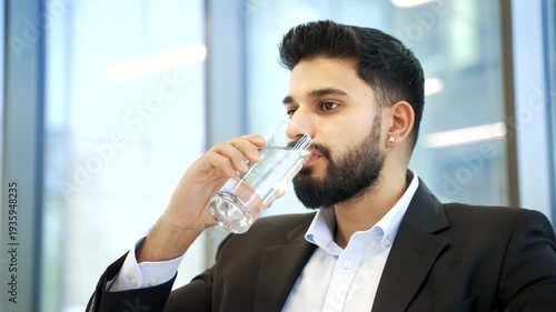 Businessman in formal suit drinks water from a glass sitting at workplace in modern business office. Calm male employee feeling relieved, enjoying clean cool drink during break at work. Close up