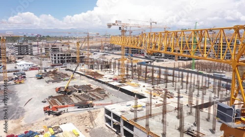 Aerial view of a massive urban construction site. Numerous yellow tower cranes and concrete building frames dominate the landscape as a large city district develops under sunny skies.