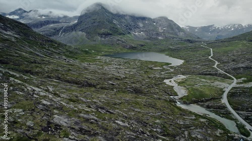 Cinematic drone pull back shot of a lonely winding road through the rugged Nordic landscape near Alnesvatnet lake, Trollstigen, Norway. Majestic mountains, green tundra and wild nature
