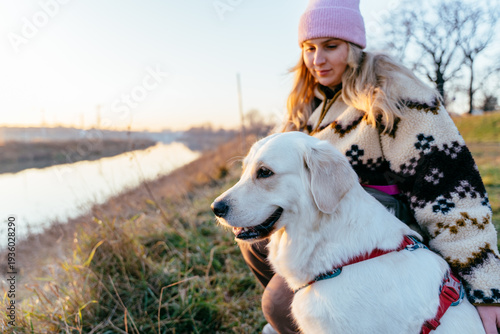 Cheerful female, blonde Caucasian, crouching with golden retriever on riverbank at sunrise