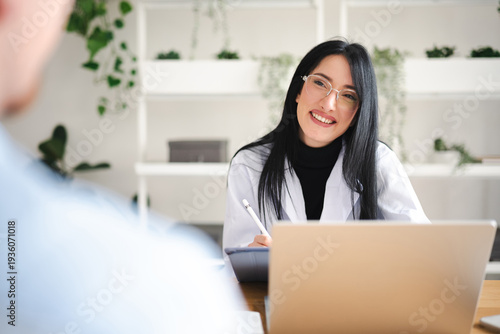 A smiling woman in a white coat and glasses is working on a laptop and tablet, interacting with a patient in a bright, modern office setting.