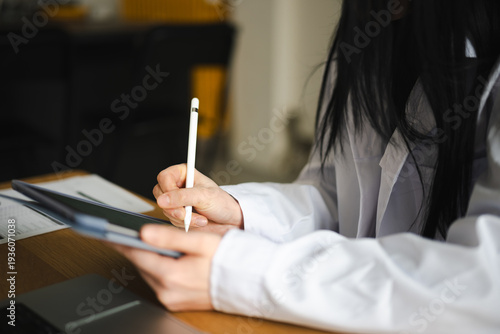 A person in a white shirt uses a stylus to write on a tablet, with papers and a laptop on the desk.