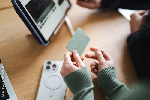 A person holds a payment card while looking at a tablet, with a smartphone and laptop nearby, suggesting online transactions or financial management.