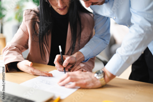 A man and woman collaborate at a desk, with the woman signing a document while the man offers guidance.