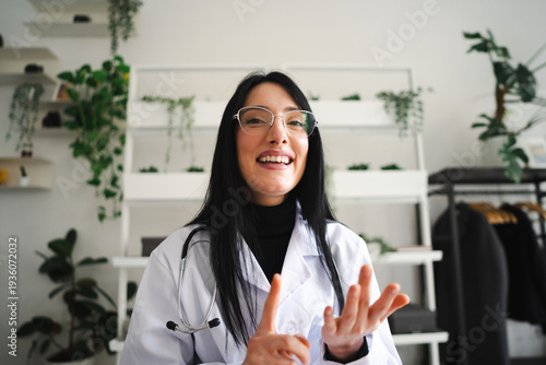 A smiling female doctor in a white coat and stethoscope gestures with her hands while speaking to the camera in a bright, modern office setting.