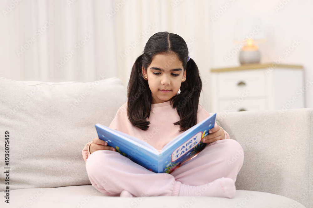 Naklejka premium Cute little girl reading book on couch at home