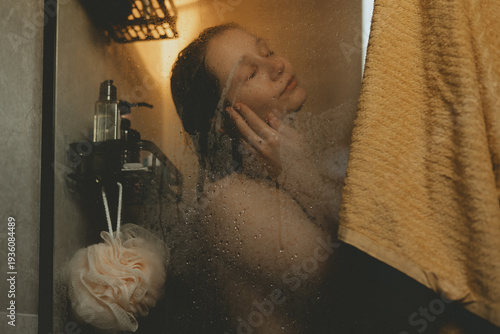 Foto Young woman practicing self-care ritual in cozy bathroom, washing hair behind glass shower door with water drops