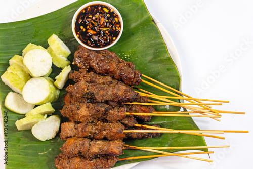Closeup of chicken satay, slices of rice cake and soy chili sauce serve with banana leaf on white background top view