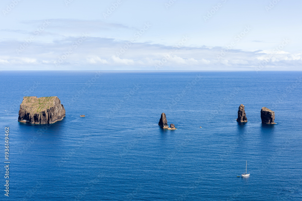 Fototapeta premium Rock formations in Atlantic Ocean off San Miguel Island coast, Azores, Portugal. Jagged rocks stand tall in blue water. Small sailboat passing by in background.